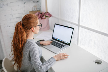 Young beautiful woman with red hair, wearing glasses, working in the office, uses a laptop and mobile phone