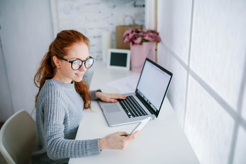 Young beautiful woman with red hair, wearing glasses, working in the office, uses a laptop and mobile phone