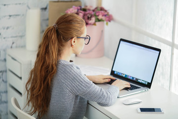 Young beautiful woman with red hair, wearing glasses, working in the office, uses a laptop and mobile phone