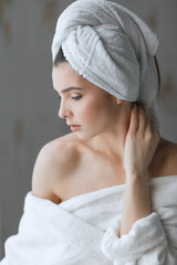 Beautiful caucasian woman with dark eyes and healthy white skin, dressed in bathrobe with naked shoulders and towel on head, nude makeup, beauty cosmetic salon atmosphere and facial spa treatment.