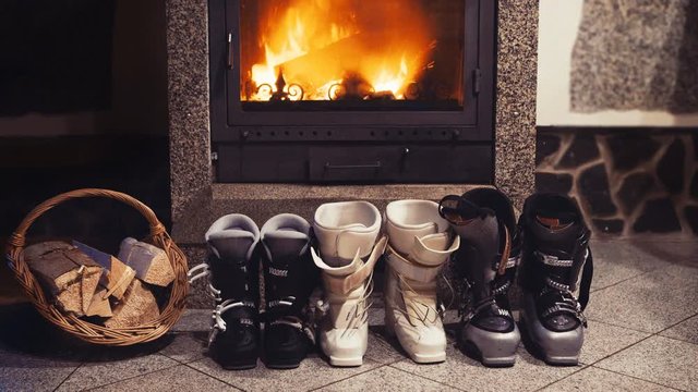 4k three pairs of skiing boots sit near glowing fireplace and basket of kindling. Winter holidays concept.