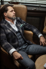 Portrait of fashionable trendy elegant wealthy professional trendsetter mature designer, dressed in tailored plaid jacket, sitting in luxury armchair in living room at home.