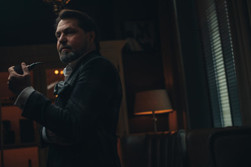 Pensive mature bearded businessman in expensive stylish suit and white shirt, no tie, smoking pipe in dark interior with dim lighting, thinking about forthcoming important meeting with partners