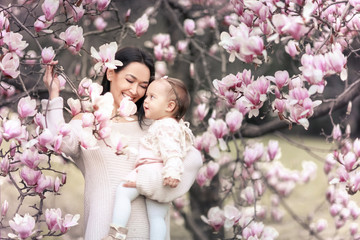 Obraz premium Cute Baby 6 month old Girl in Pink Outfit with Big Blue Eyes with Young Beautiful Mother at Spring, Pink Blooming Tree at the Background
