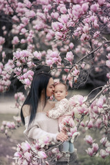 Fototapeta premium Cute Baby 6 month old Girl in Pink Outfit with Big Blue Eyes with Young Beautiful Mother at Spring, Pink Blooming Tree at the Background