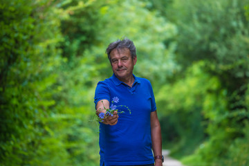 Beautiful matured men in blue teeshirt on the rural road in the summer forest giving blue wildfllowers