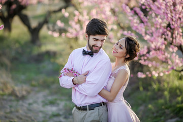 Newlyweds in love stand on the nature, against the background of wooden stakes, in sunny weather. Stylish groom embraces a beautiful bride in a lace dress in a green garden.