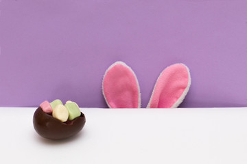 Chocolate egg filled with candy and bunny ears peeking from the table