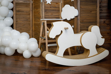 Fototapeta na wymiar Birthday white decorations with balloons, stars, clouds and wooden horse for little baby on a dark brown background.