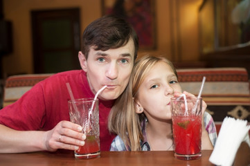 Young girl of adolescence along with her dad have fun at the cafe - drinking crimson juice with ice through a tube
