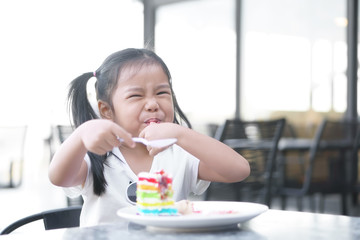 Asian child cute or kid girl smiling enjoy and fun with happy eating delicious rainbow cake and suck finger for sweet and sour dessert on table and holding spoon at lunch in cafe restaurant or shop