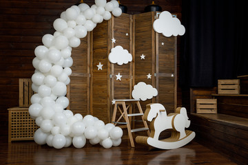 Birthday white decorations with balloons, stars, clouds and wooden horse for little baby on a dark brown background.