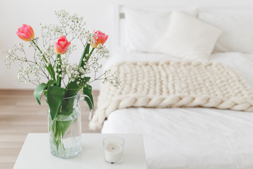 Glass vase with pink tulips and gypsophila flowers in light cozy bedroom interior. White wall, bed with white linen, beige thick yarn knitted woolen merino chunky blanket or plaid, pillows, candle.