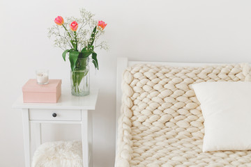 Cozy bedroom interior: white wall, bed with white linen, light beige thick yarn knitted woolen merino chunky blanket or plaid, pillows, bedside table, glass vase with tulips flowers.