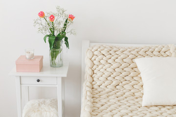 Cozy bedroom interior: white wall, bed with white linen, light beige thick yarn knitted woolen merino chunky blanket or plaid, pillows, bedside table, glass vase with tulips flowers.