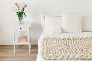 Cozy bedroom interior: white wall, bed with white linen, light beige thick yarn knitted woolen merino chunky blanket or plaid, pillows, bedside table, vase with tulips flowers.
