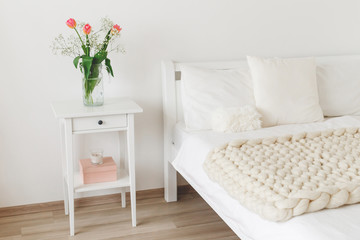 Naklejka premium Cozy bedroom interior: white wall, bed with white linen, light beige thick yarn knitted woolen merino chunky blanket or plaid, pillows, bedside table, vase with tulips flowers.