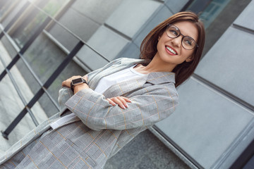 Business casual. Young lady in eyeglasses standing on the city street crossed arms looking aside happy