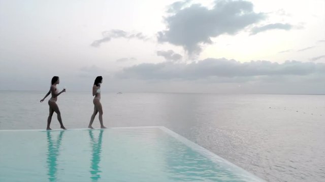 Aerial: Two Sexy Young Women Walking on the Edge of Swimming Pool by the Ocean