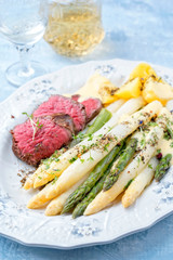 Traditional white and green asparagus with barbecue dry aged sliced beef fillet and fried potatoes served as closeup on a shabby chic design plate