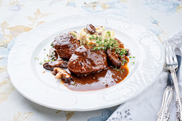 Traditional German braised pork cheeks in brown red wine sauce with mushroom and mashed potatoes as closeup on a classic design white plate