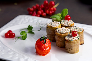 Eggplant rolls with a filling, on a white plate of urshenoy baked tomato. Dark background decorated with vegetables and viburnum