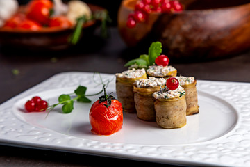 Eggplant rolls with a filling, on a white plate of urshenoy baked tomato. Dark background decorated with vegetables and viburnum
