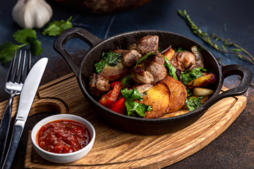Frying pan with roast beef and potatoes, roasted vegetables and greens, on a light board on a black background. The background is decorated with vegetables.