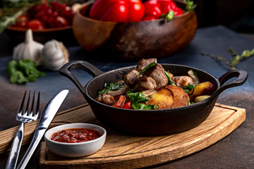 Fototapeta na wymiar Frying pan with roast beef and potatoes, roasted vegetables and greens, on a light board on a black background. The background is decorated with vegetables.