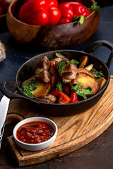 Frying pan with roast beef and potatoes, roasted vegetables and greens, on a light board on a black background. The background is decorated with vegetables.