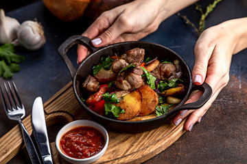 Frying pan with roast beef and potatoes, roasted vegetables and greens, on a light board on a black background. The background is decorated with vegetables.