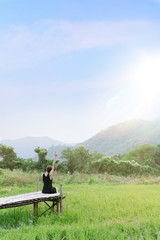 Fototapeta na wymiar A woman with black dress sitting on a the wooden bridge and raise her hand and making the sign “I love you” on the fresh green field with mountains, blue sky and sunlight flare.