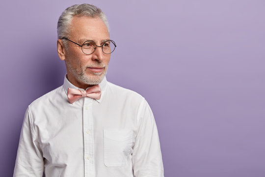 Sideways shot of thoughtful grey haired man with thick beard, wears spectacles, elegant shirt, focused aside, deep in thoughts, thinks up next step, isolated over purple studio wall with empty space