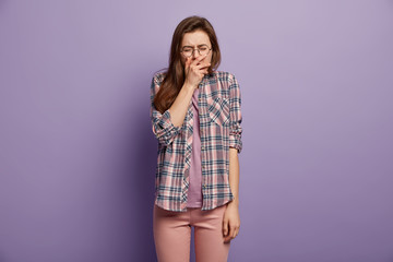 Sad woman in stress cries desperately, has embarrassed facial expression, expresses sadness, suffers from hurt feelings, dressed in checkered shirt, casual trousers, isolated over purple background