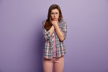 Gloomy stressful European woman keeps both hands on neck, has thore throat or asthma, cant breath well, wears round spectacles and checkered shirt, poses over purple background. Cough attack