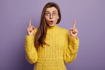Photo of astonished woman dressed in yellow jumper, wears spectacles, points up with both fore...