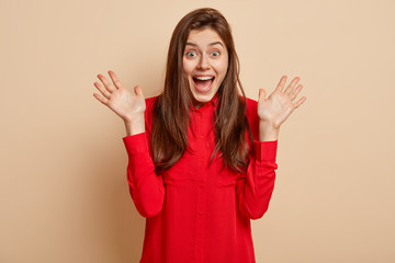 Photo of positive brunette woman spreads palms, keeps jaw dropped, dressed in red shirt, expresses positive emotions, poses over beige background. People, human facial expressions and feelings