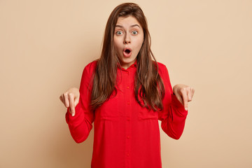 Photo of dissatisfied surprised young woman points down on floor, dressed in red shirt, isolated...