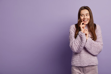Flirty young girl in winter loose sweater smiles sensually, glances aside, keeps hand near mouth, rejoices something pleasant, focused aside, isolated over purple background with empty space left