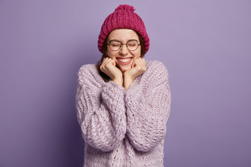 Photo of happy European girl hears positive news, keeps hands under chin, wears hat and jumper,...