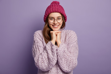 Studio shot of pleased smiling lady keeps hands together under chin, wears warm hat and loose sweater, isolated over purple background. People, good emmotions, feelings concept. Woman ready for winter