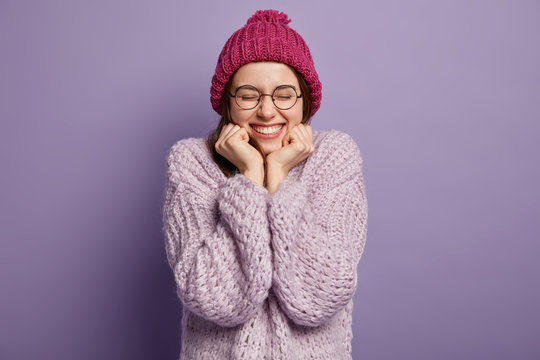 Photo of happy European girl hears positive news, keeps hands under chin, wears hat and jumper, smiles broadly, rejoices good winter day, isolated over purple background. Cheering energetic woman