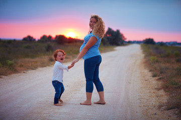 happy pregnant mother and toddler baby boy walking barefoot on countryside road at summer sunset