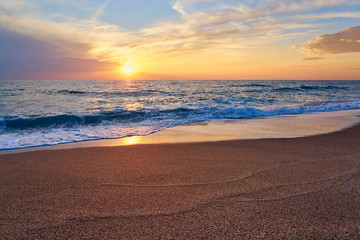 Obraz premium Colorful sunset at the tropical beach, sun behind clouds reflects on water and waves with foam hitting sand.