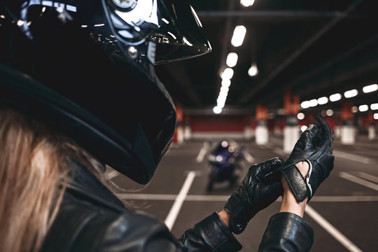 Close up image of female hands in leather gloves. Unrecognizable woman with blonde hair standing alone on car parking with empty spaces, wearing safety helmet and black jacket. Film effect