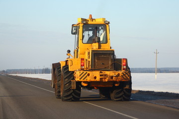 Heavy power yellow bulldozer tractor rides along the asphalt road on the background of snowy fields and the blue of a clear sky in winter - rear side view