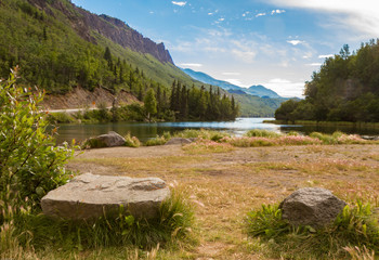 Two rocks on ground in front of view of lake, forest, and mountains in Alaska, USA. Rocks in foreground, lake in mid ground, mountains and trees on left of frame.