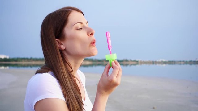 Happy young woman having fun outdoor. Cheerful female blowing soap bubbles on lake at dusk in slow motion. Concept of freedom and happiness