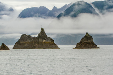 Two pinnacle rock formations in Aialik Bay, Alaska, USA. Rock formations are surrounded by white mist with mountains in the background. 