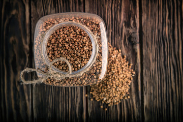 Bank with buckwheat on wooden background. A bunch of buckwheat lies next to the can. View from above.